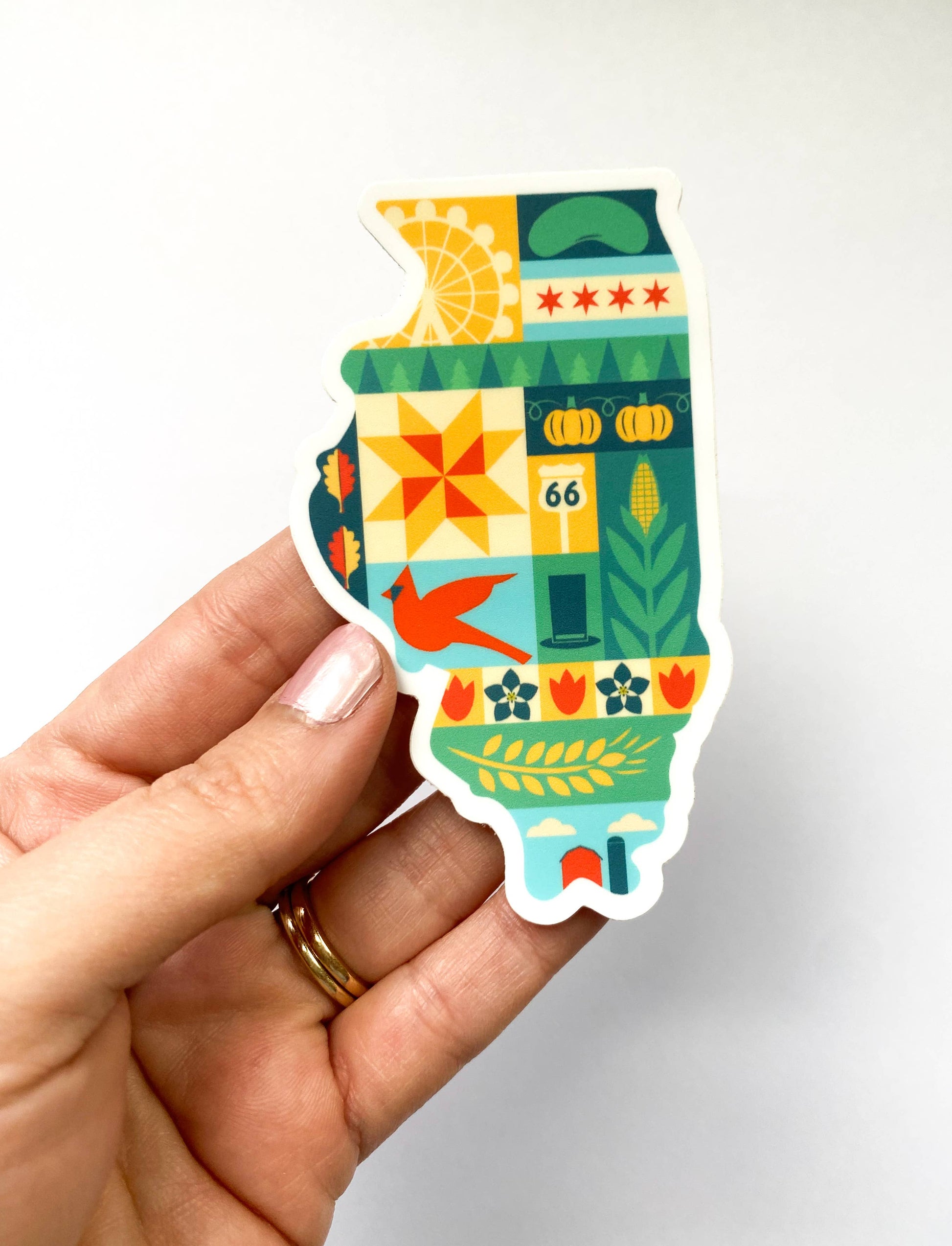 A hand holding a die-cut sticker shaped like the state of Illinois, featuring a colorful retro-style patchwork design with farm, city, and decorative motifs on a plain light background.