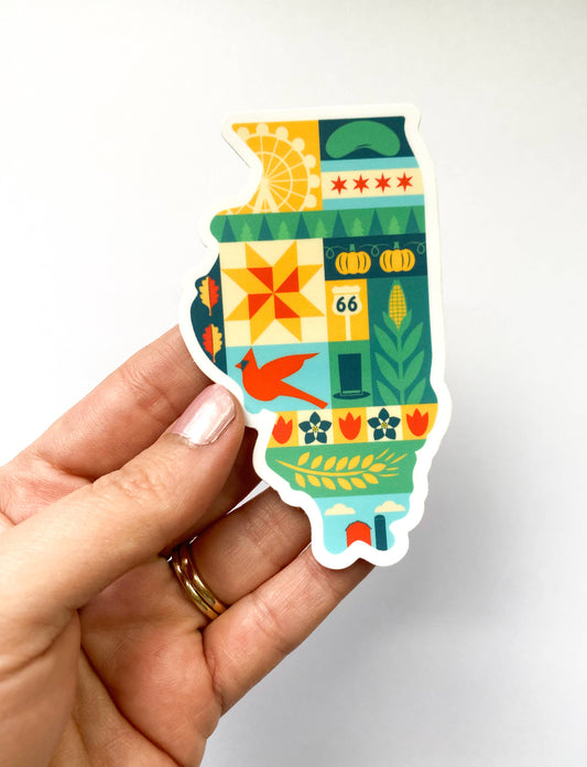 A hand holding a die-cut sticker shaped like the state of Illinois, featuring a colorful retro-style patchwork design with farm, city, and decorative motifs on a plain light background.