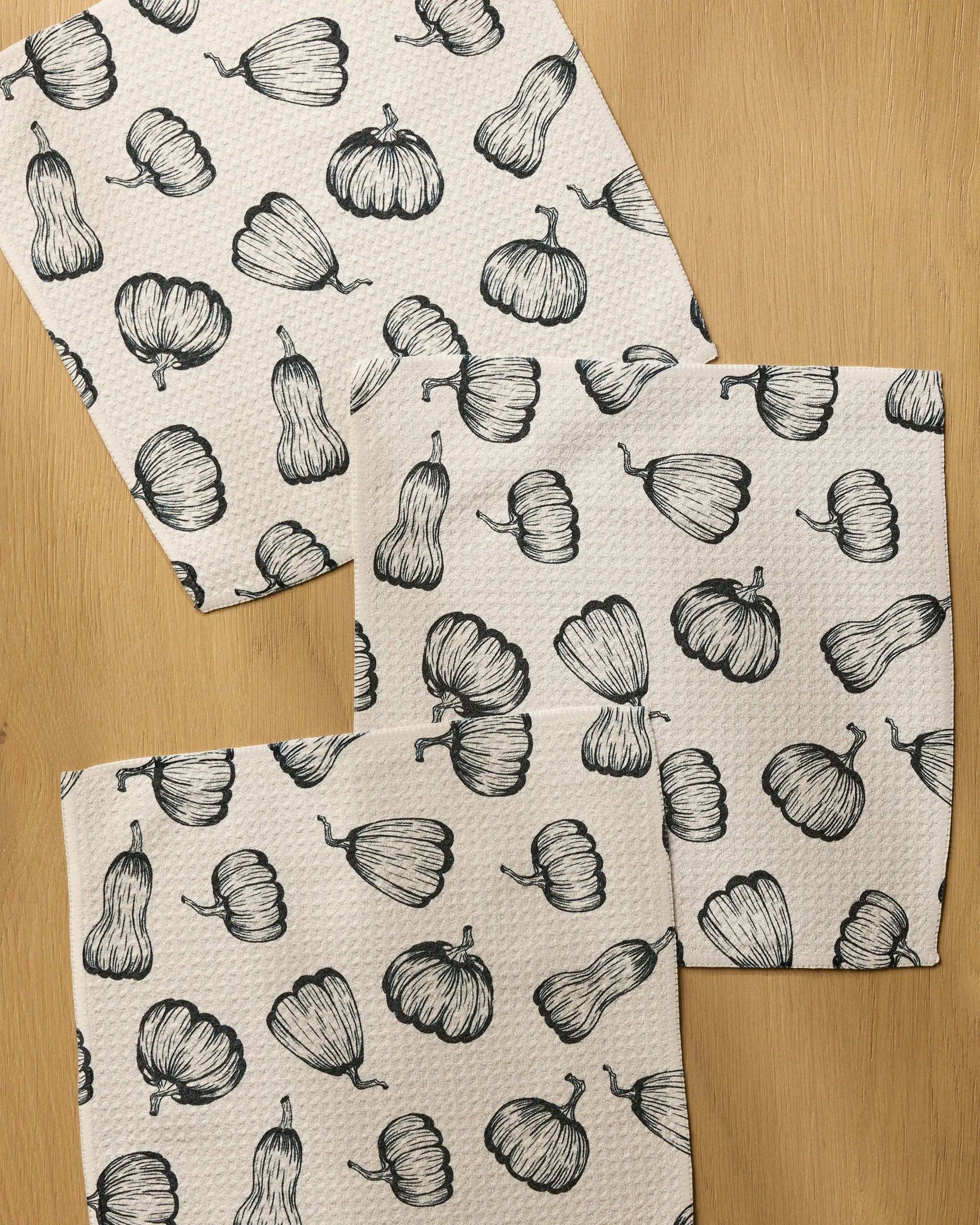 Three square kitchen towels with a black and white pumpkin pattern, arranged on a light wood surface.
