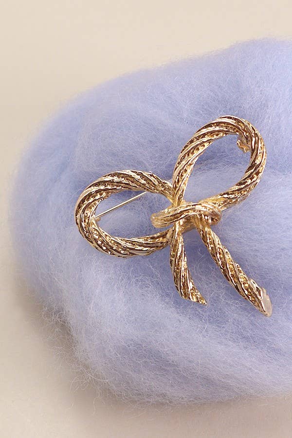 Gold bow shaped bow brooch with rope texture, resting on a purple cotton tuft