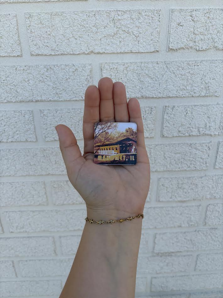 A hand holding a small square printed card or magnet featuring a scenic image of a yellow building and autumn trees, set against a white brick wall background.