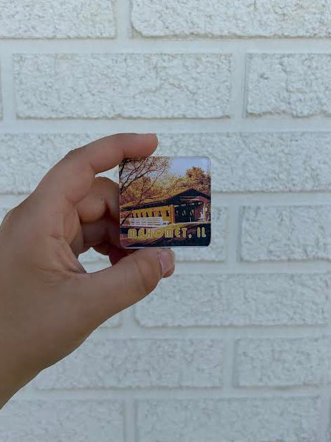 A hand holding a small square printed card or magnet featuring a scenic building and trees, set against a white textured brick wall.