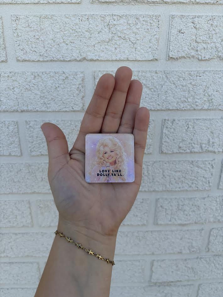 A hand holding a small square card featuring a portrait of Dolly Parton with the text 'LOVE LIKE DOLLY Y'ALL' against a white brick wall background.