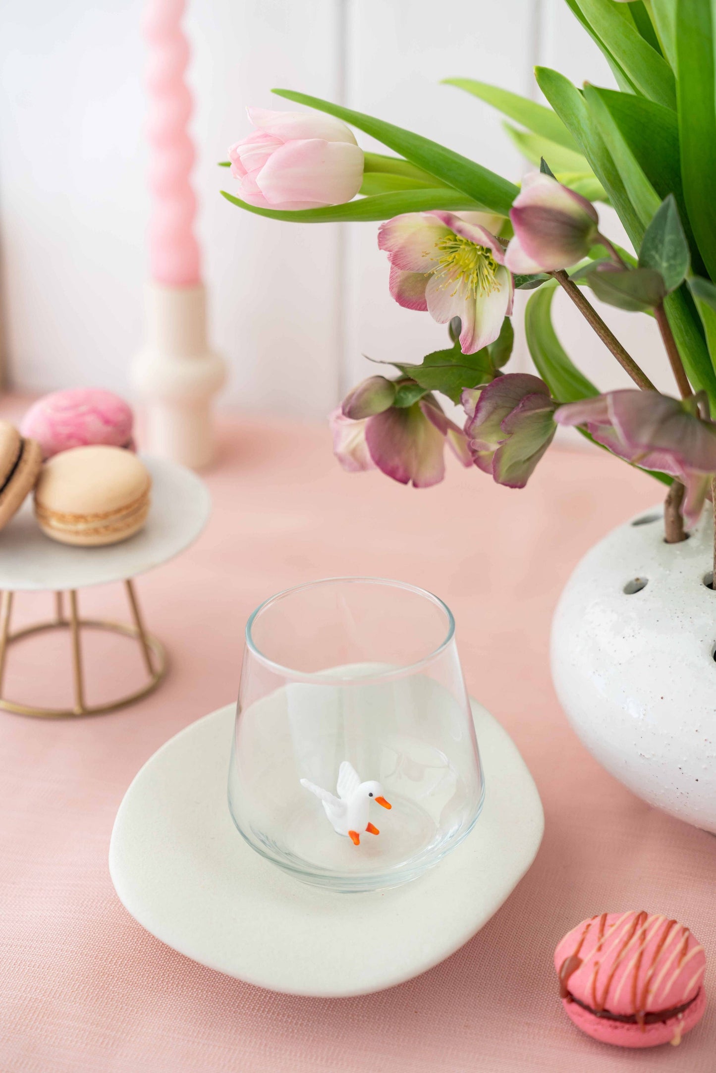 Glass with a duck figure on a pink surface with flowers and macarons.