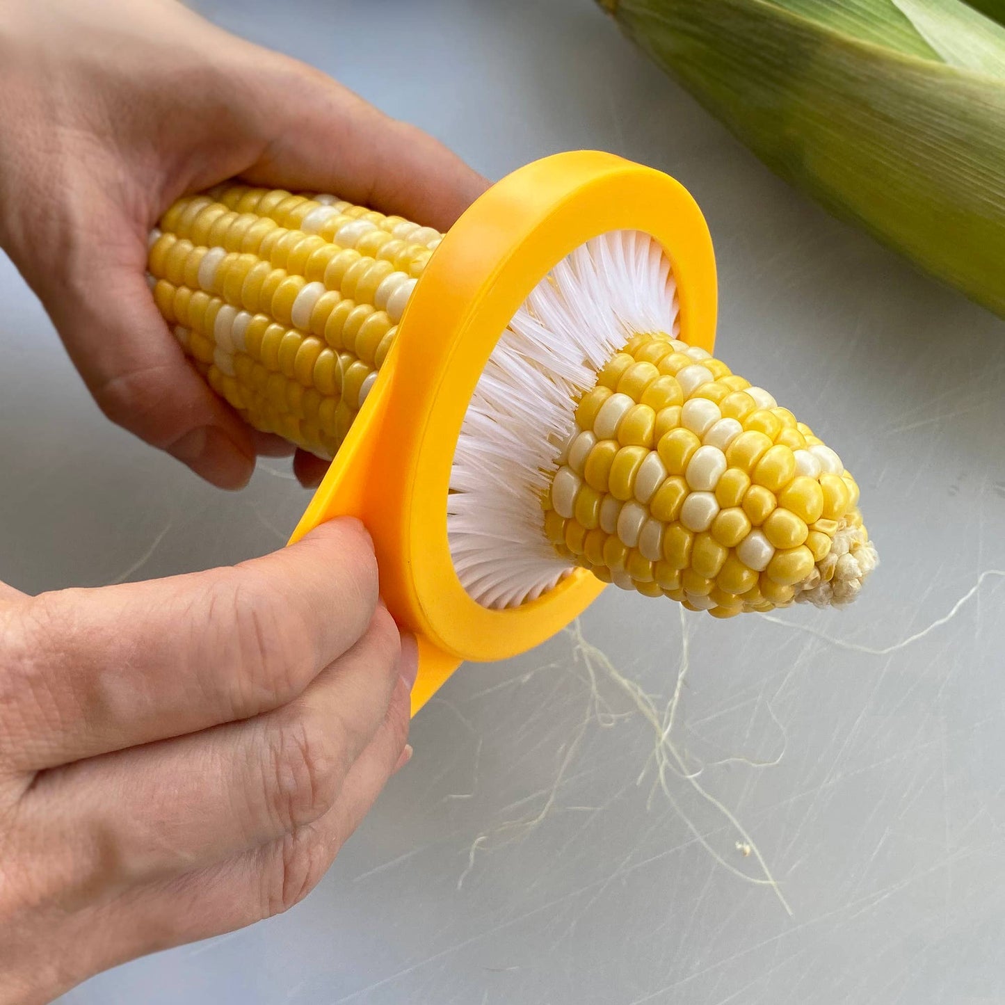 Corn on the cob being cleaned with a yellow corn cleaner tool on a white surface.