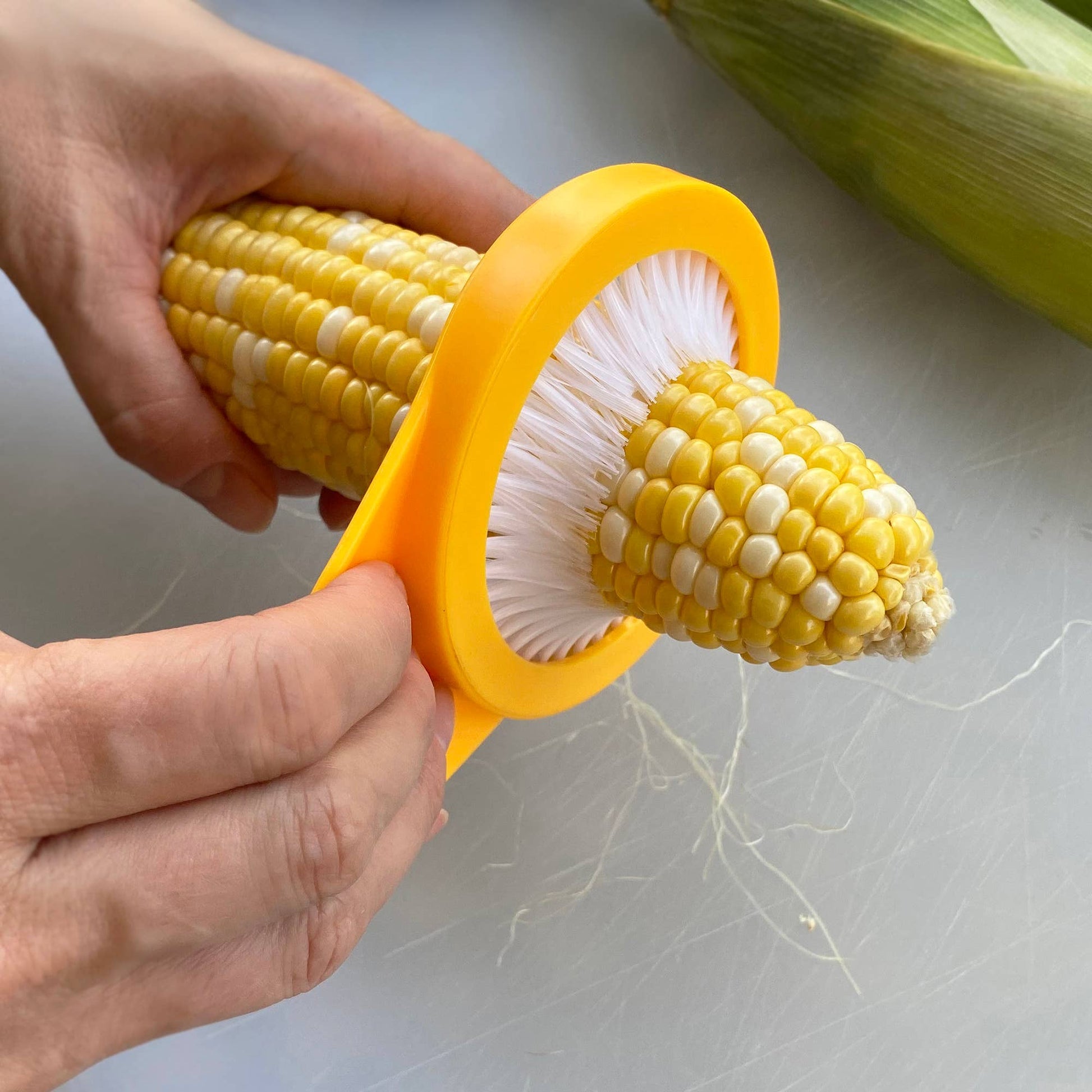 Corn on the cob being cleaned with a yellow corn cleaner tool on a white surface.