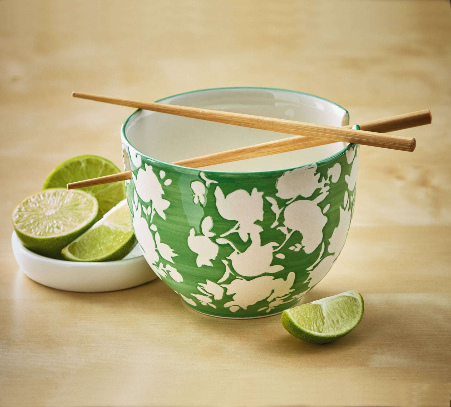 Green noodlebowl pictured with a dish of limes for scale. The chopsticks rest on top of the bowl. The background is a pale wooden texture.