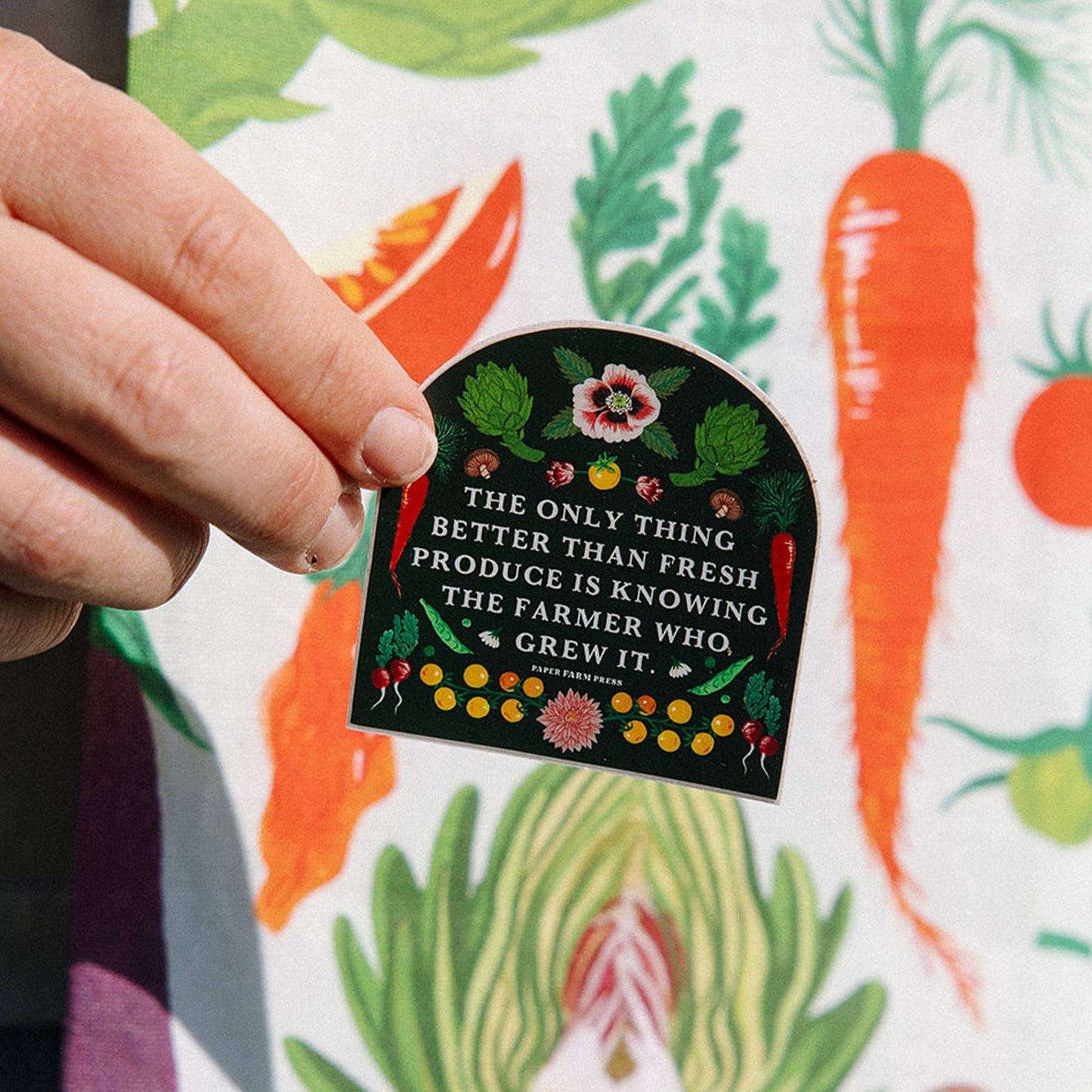 Hand holding a decorative sticker with a quote about fresh produce against a colorful background of vegetables.