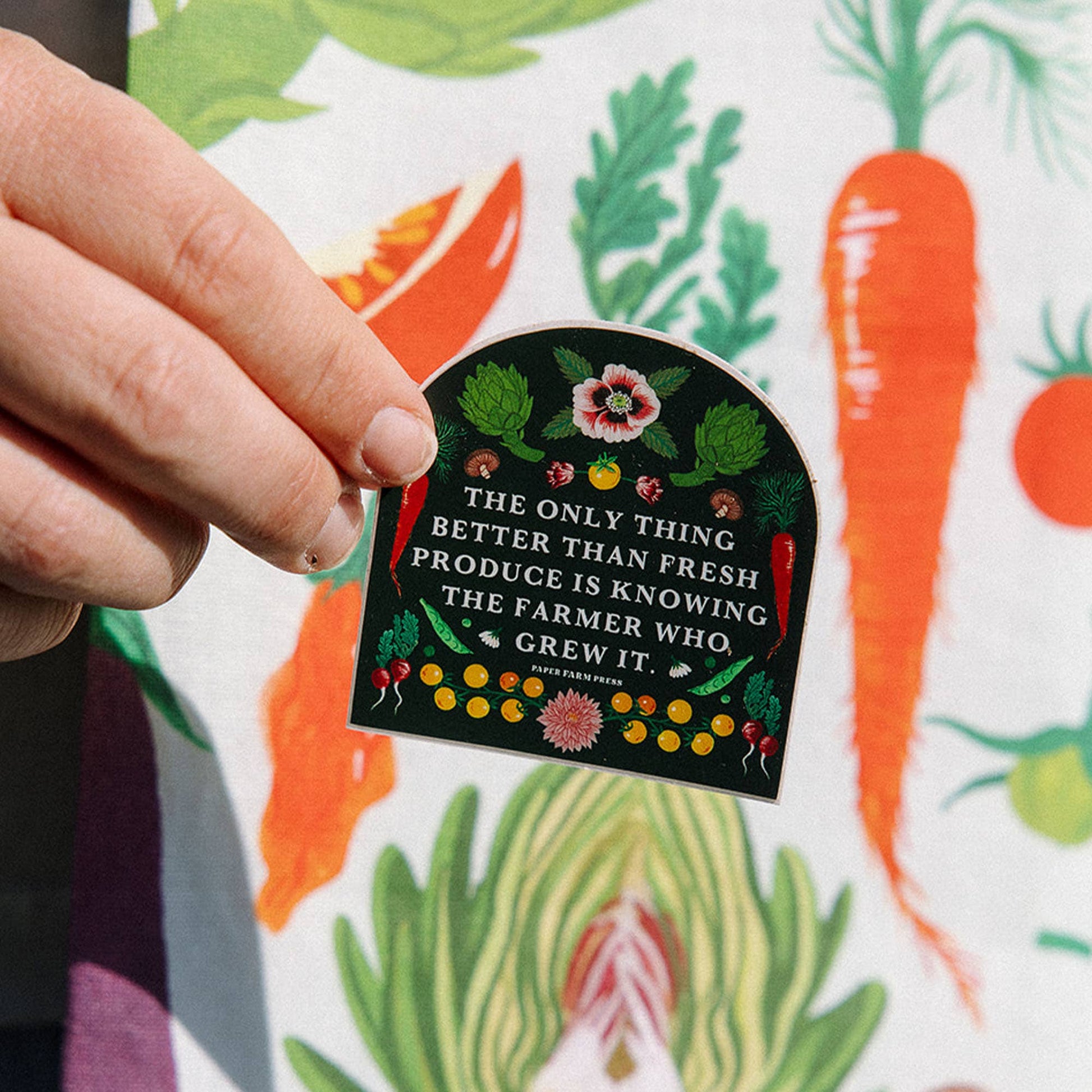 Hand holding a decorative sticker with a quote about fresh produce against a colorful background of vegetables.