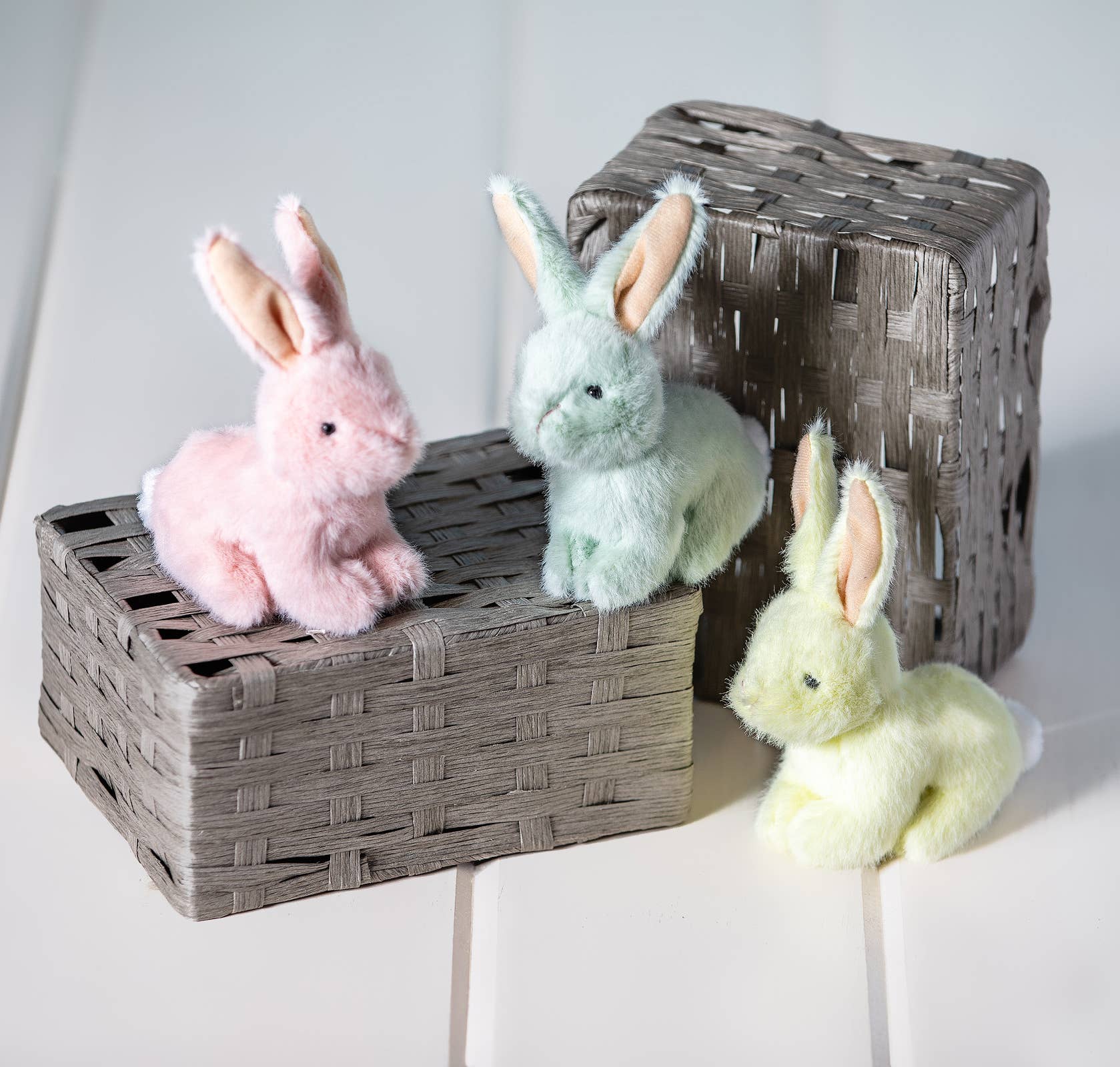 Three plush bunny toys in pink, green, and yellow on a woven basket against a white background.
