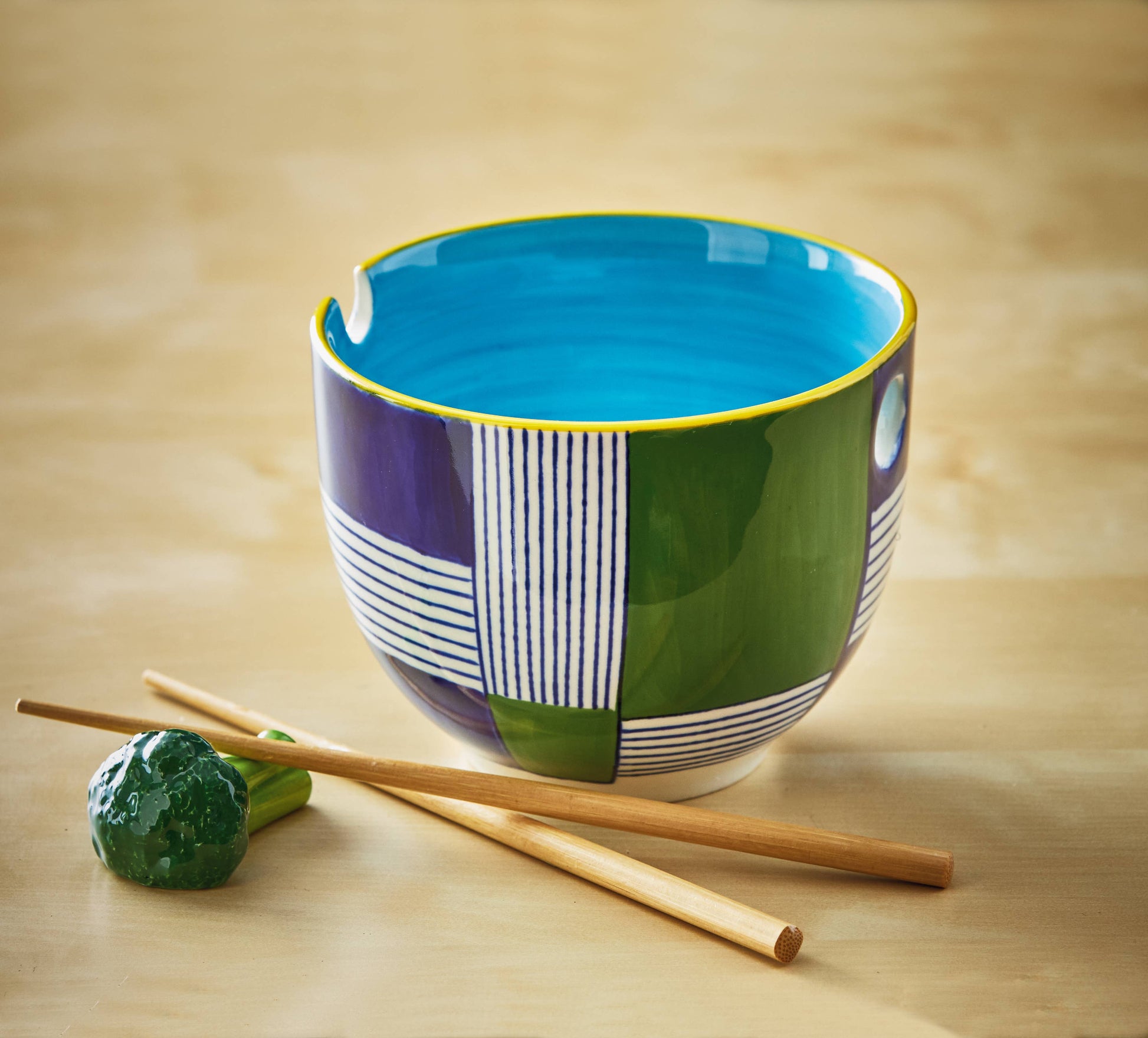 Noodle bowl is show next to a pair of chopsticks (included with the purchase of this item) and a broccoli shaped chopstick rest (not included with the purchase of this item). Everything is on a wooden background. 