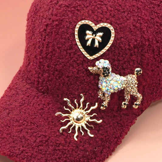 Three golden brooches, one poodle, one black heart with bow, and one sun, pinned to a red sherpa ball cap on a pink background