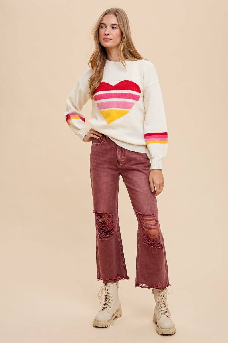 Woman wearing an ivory sweater with heart motif in the center made of 4 red, hot pink, light pink, and yellow stripes, and stripes of the same colors around the sleeves