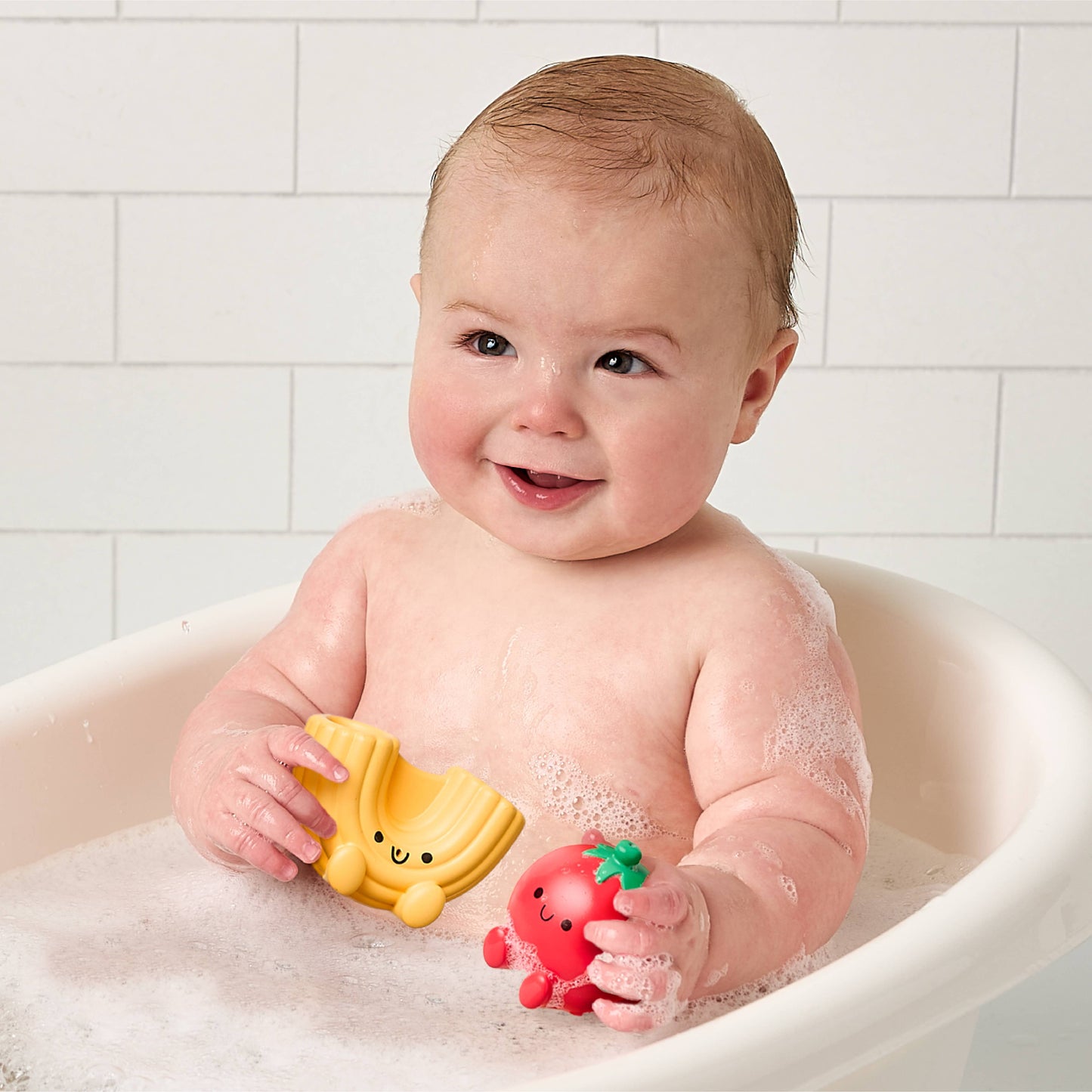 Baby in a bathtub holding two bath toys, one macaroni noodle and one tomato, against a tiled bathroom wall.