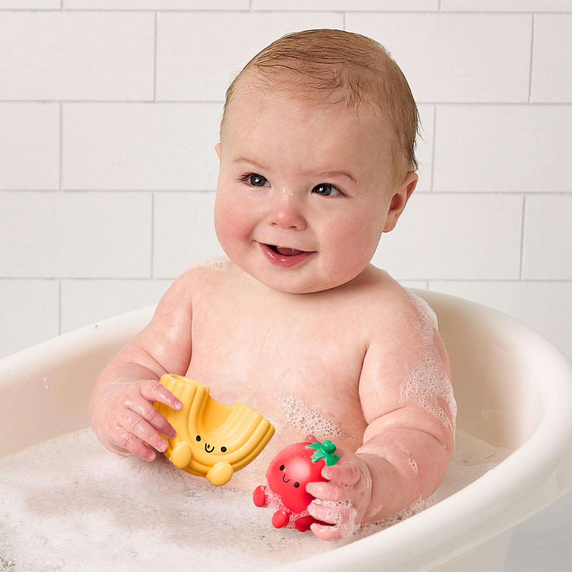 Baby in a bathtub holding two bath toys, one macaroni noodle and one tomato, against a tiled bathroom wall.