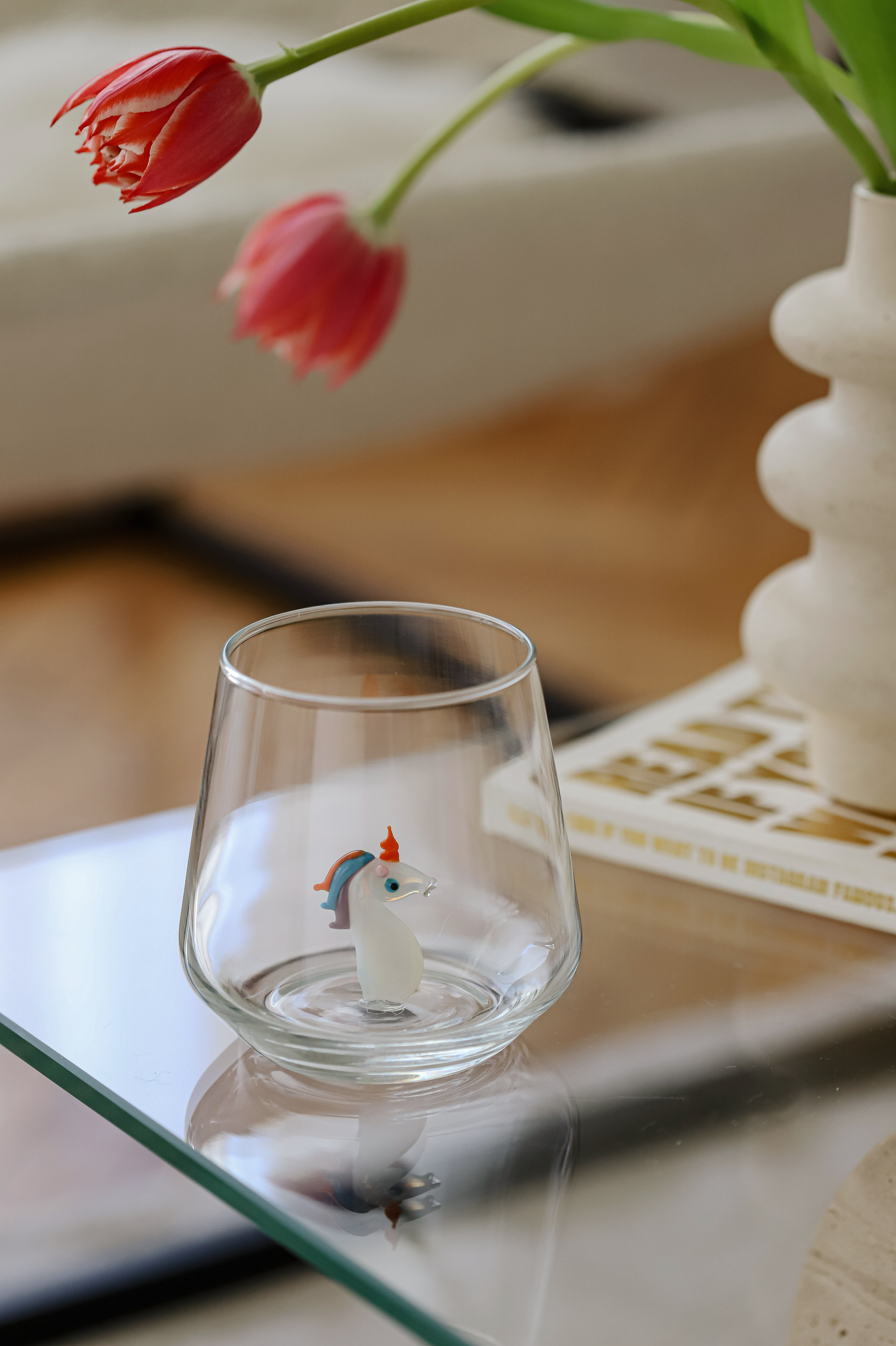 Clear glass with a small figurine on a reflective surface, with a blurred background of a vase and flowers.