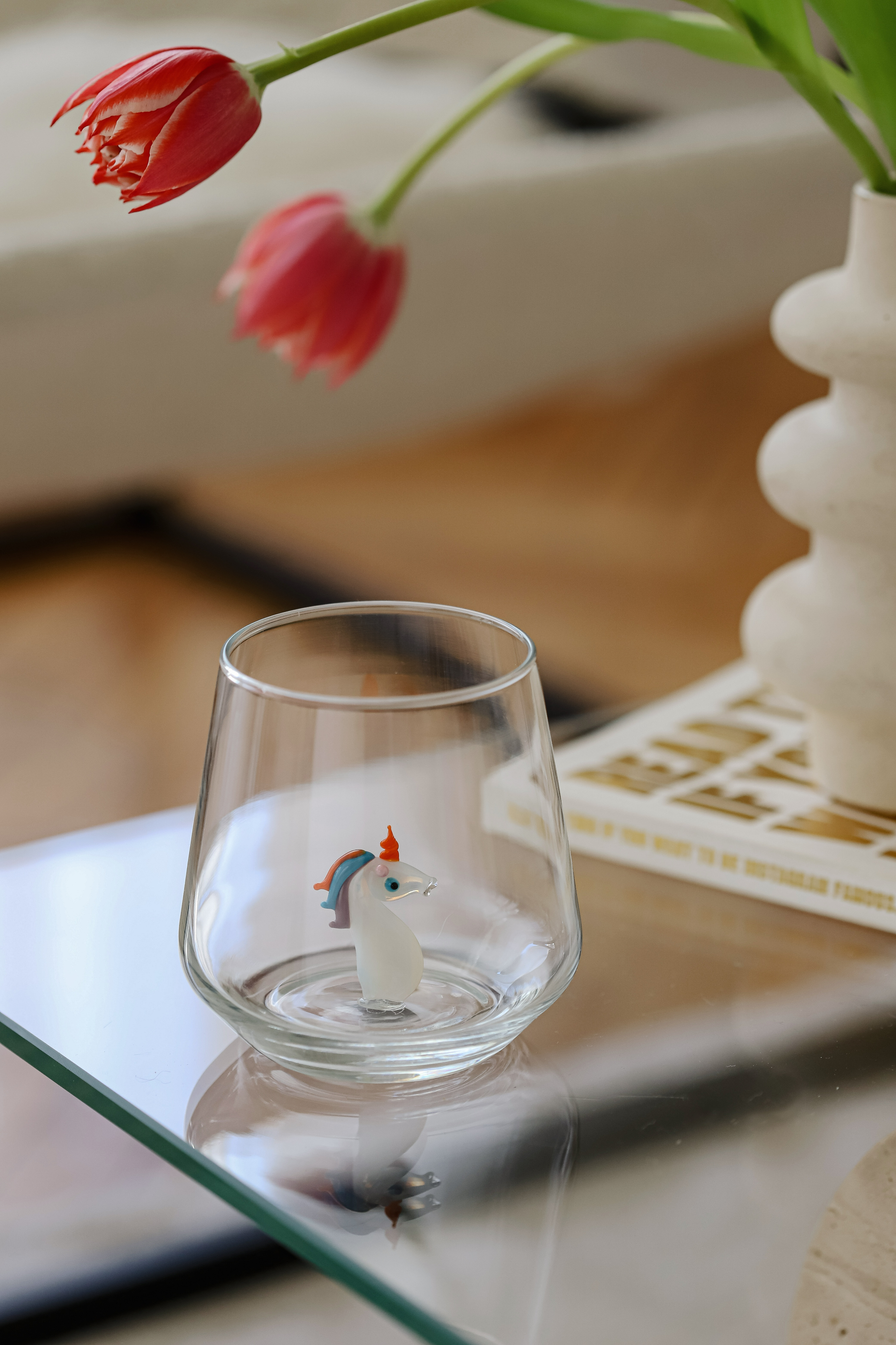 Clear glass with a small figurine on a reflective surface, with a blurred background of a vase and flowers.