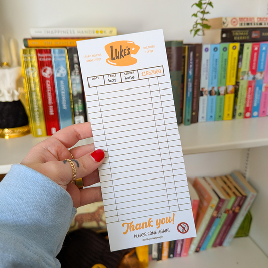 Hand holding a notepad with 'Luke's' branding in front of a bookshelf.