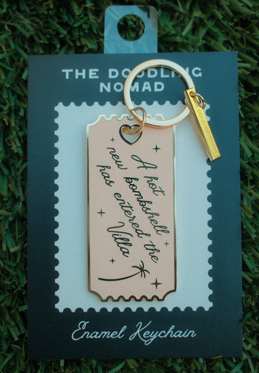 A rectangular enamel keychain with a gold-tone split ring and small gold tag, displayed on a branded backing card. The keychain has a pale pink enamel body with black script text reading 'A hot beach ball has entered the Valley' and small black decorative marks.