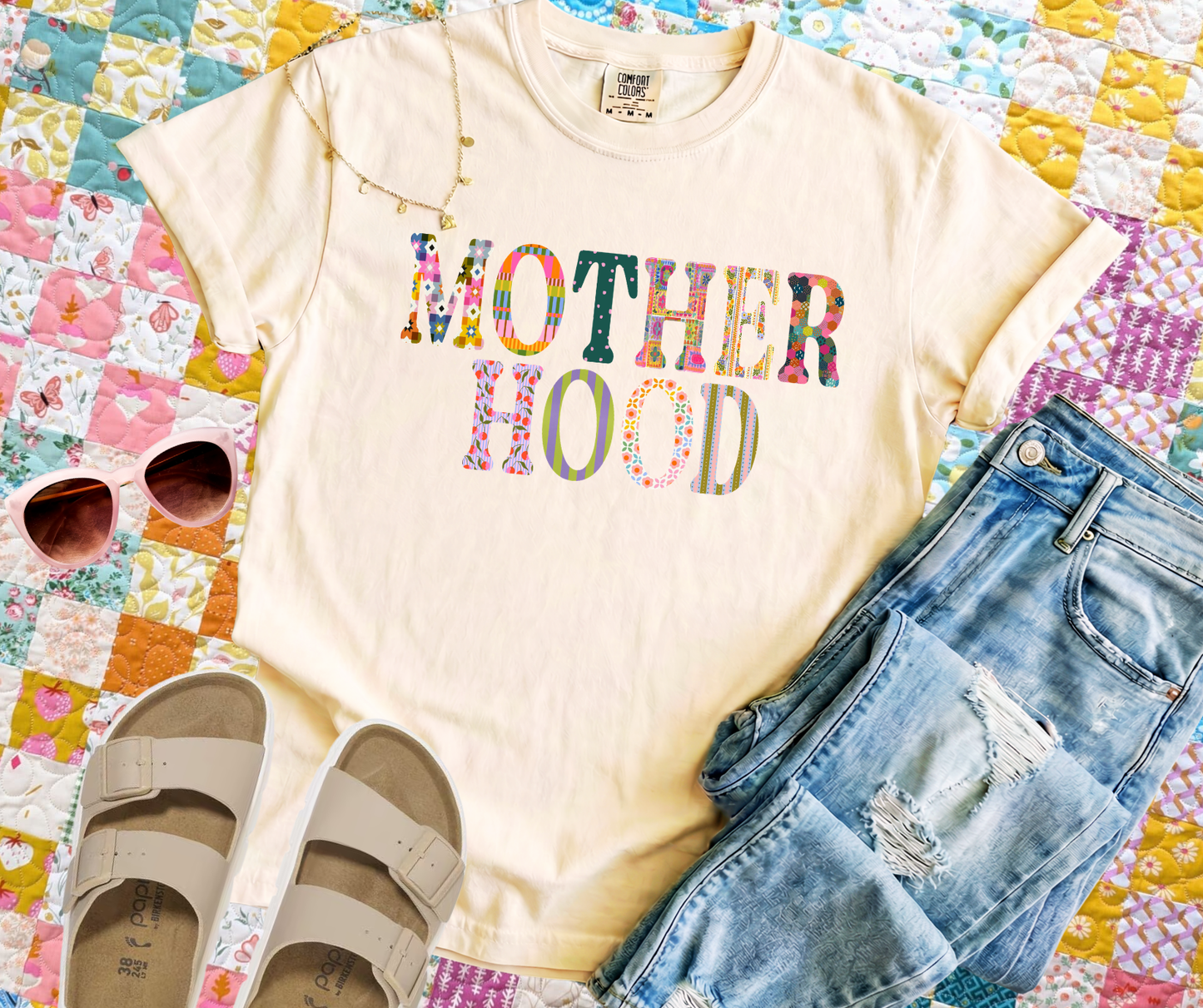 T-shirt with 'MOTHERHOOD' text, sunglasses, and denim shorts on a colorful quilt background