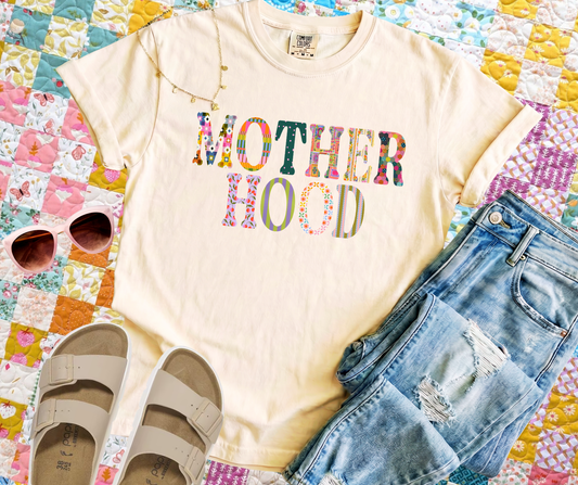 T-shirt with 'MOTHERHOOD' text, sunglasses, and denim shorts on a colorful quilt background