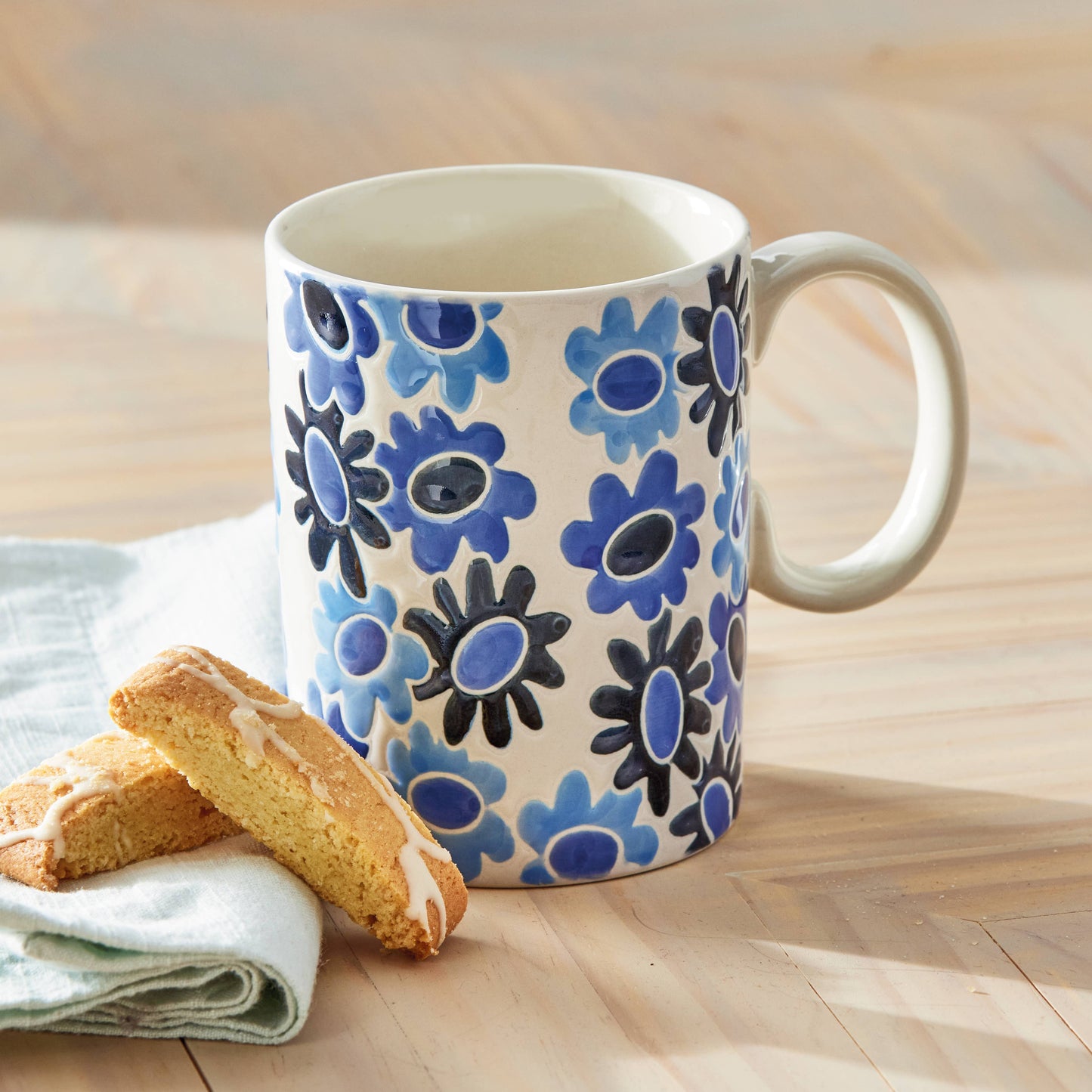 White mug with blue 'mod' styled flowers design sitting on a wooden countertop with a biscotti and cloth napkin beside it.