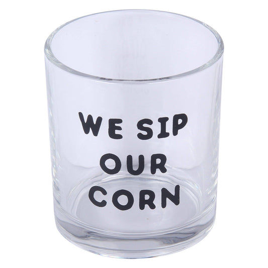 A clear glass tumbler with the humorous phrase 'WE SIP OUR CORN' printed in black capital letters on the front, set against a plain white background.