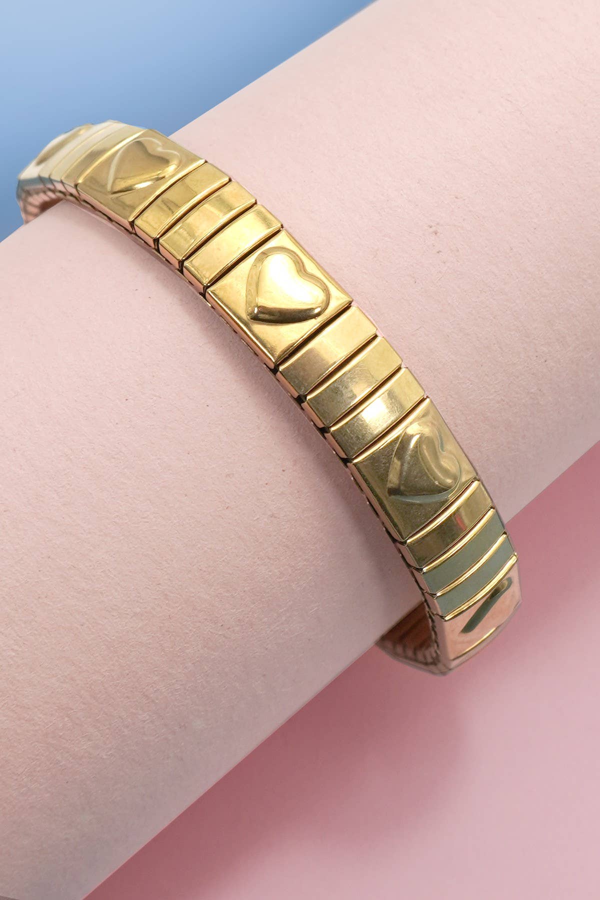 Gold bracelet with heart design on a wrist against a pink and blue background
