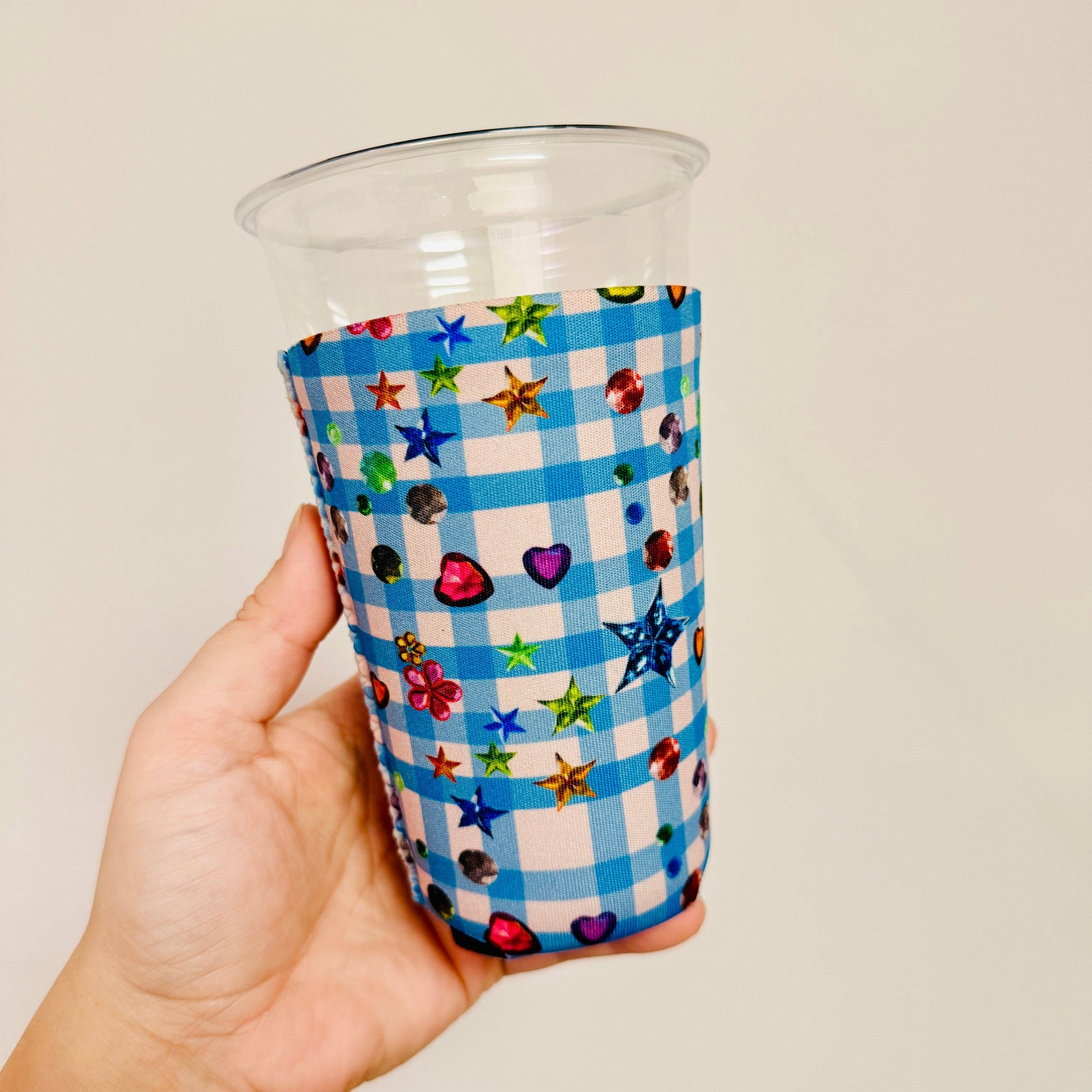 Hand holding a clear plastic cup with a blue checkered sleeve featuring colorful stars and hearts.