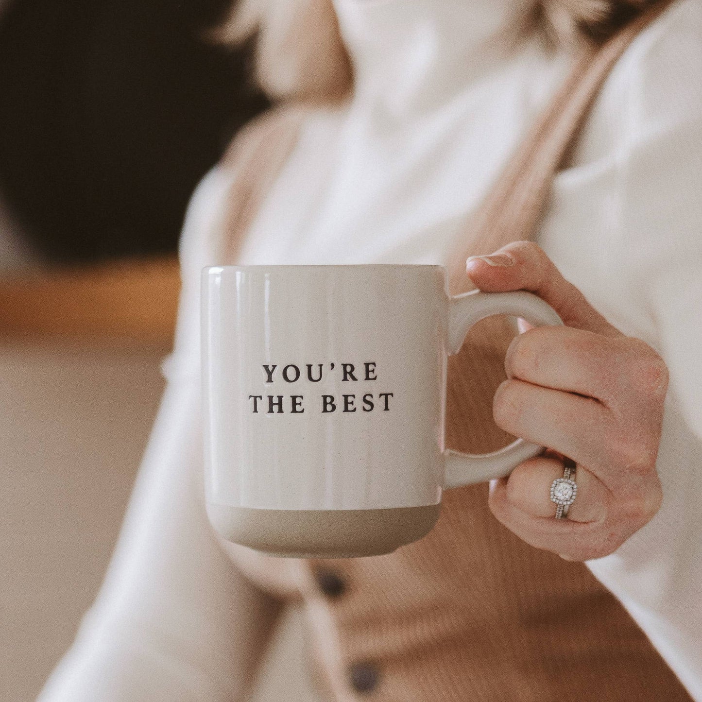 Person holding a mug with 'YOU'RE THE BEST' text, wearing a beige sweater.