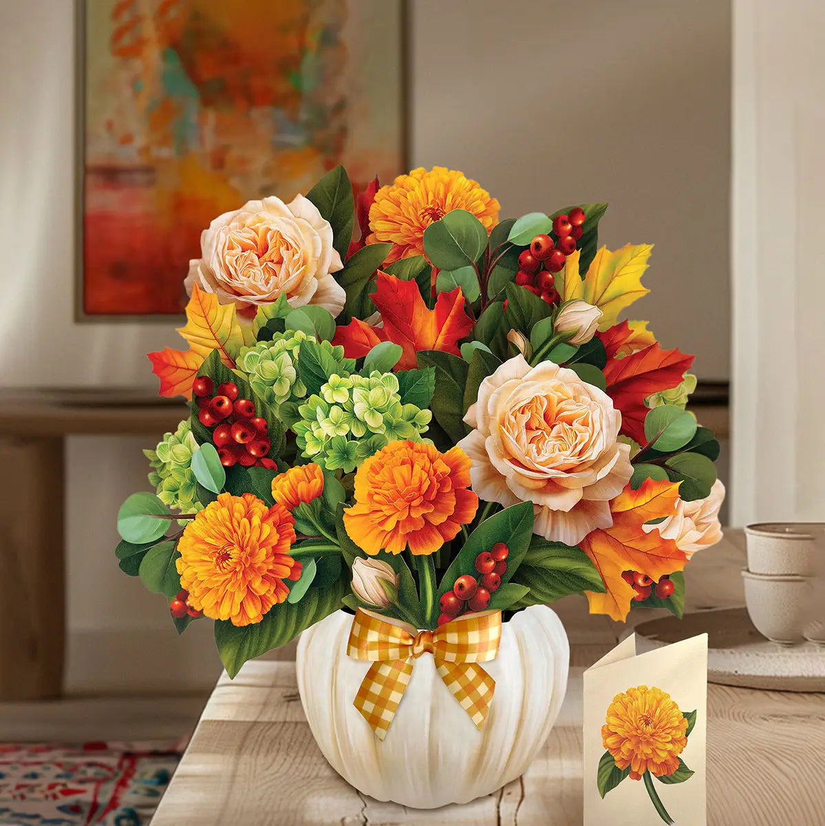 A decorative autumn floral arrangement in a white pumpkin-shaped vase with a yellow gingham bow, placed on a wooden table in a home interior. The bouquet includes orange marigolds, peach roses, red berries, green hydrangeas, and fall leaves.