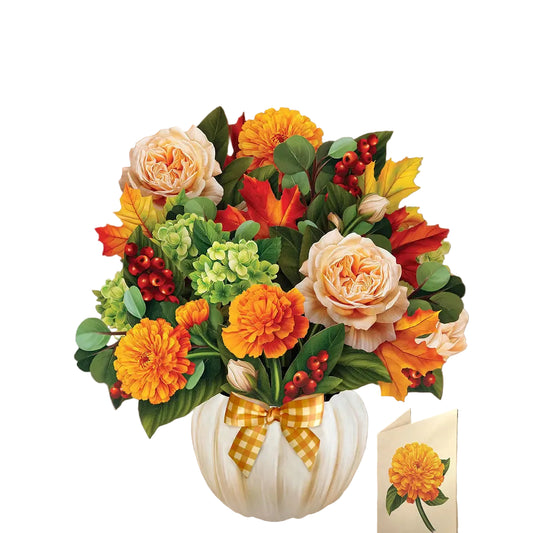 A decorative autumn floral arrangement in a white pumpkin-shaped vase with a yellow gingham bow, placed on a wooden table in a home interior. The bouquet includes orange marigolds, peach roses, red berries, green hydrangeas, and fall leaves.