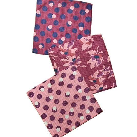 Three square fabric napkins with pink, purple, and blue patterns, including polka dots and floral motifs, arranged on a light wood background.