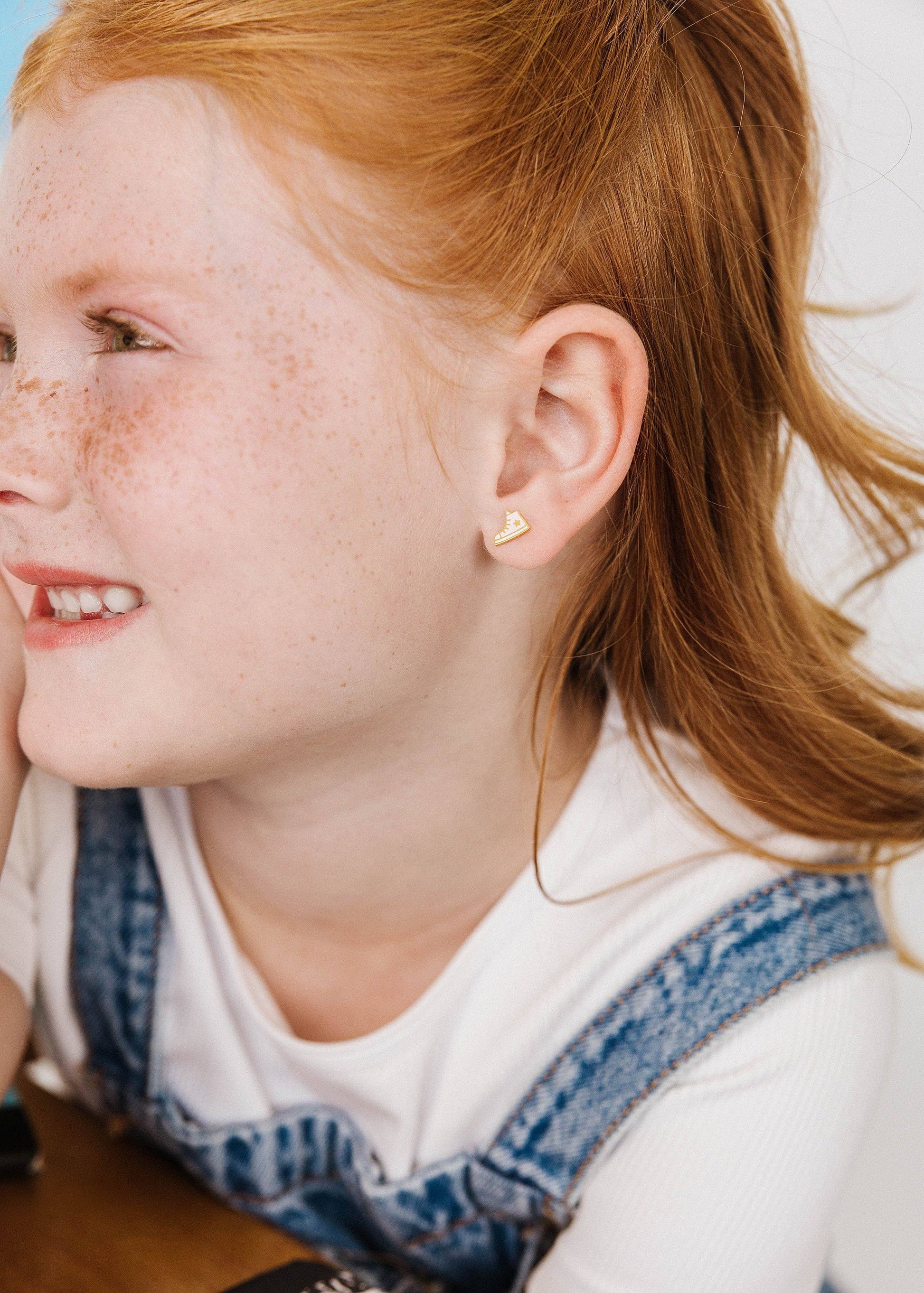 A close-up side profile of a young girl with red hair and freckles, wearing a high-top sneaker shaped stud earring, a white t-shirt, and blue denim overalls.