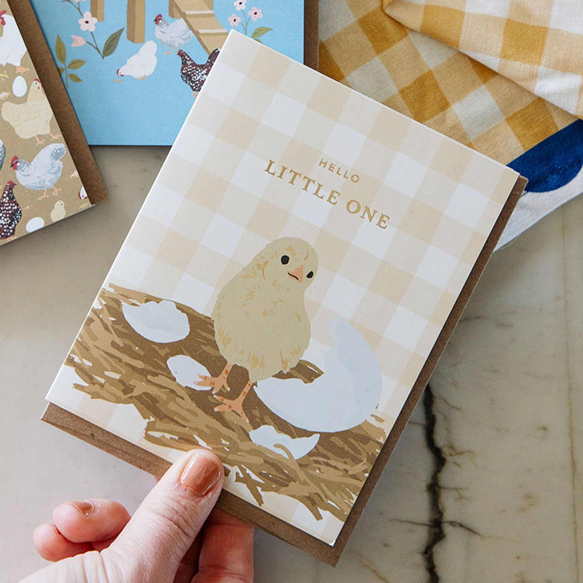 Card with a chick illustration and 'Hello Little One' text, held by a hand on a marble surface.