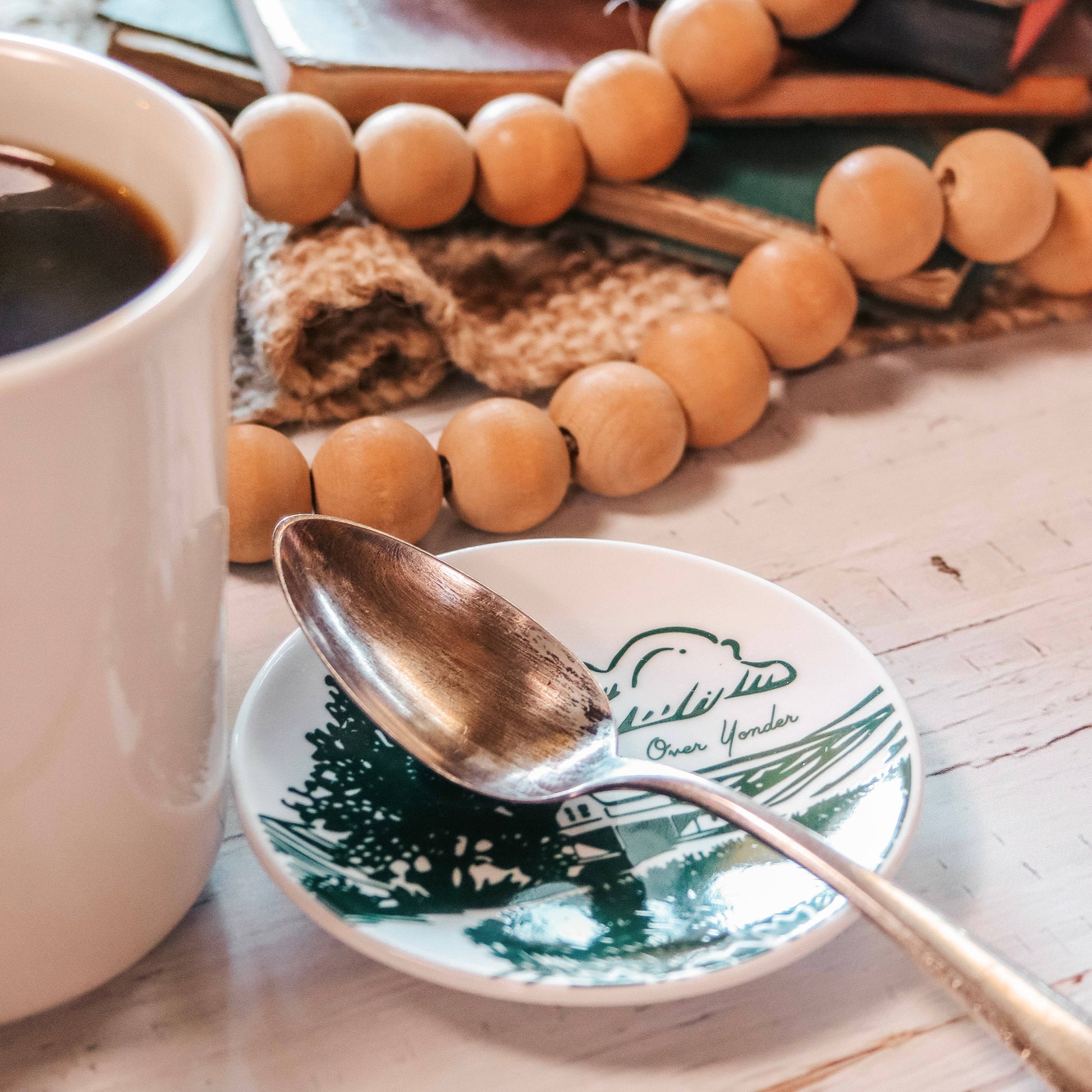 Cup of coffee with a spoon on a decorative plate, surrounded by wooden beads and a knitted item.