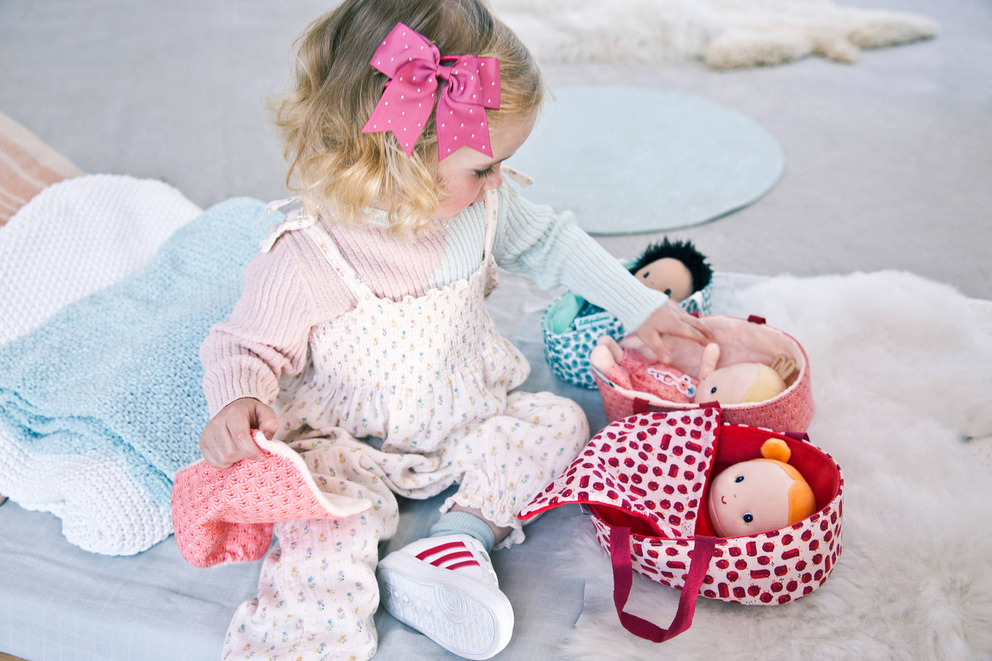 Child playing with toys on a bed, wearing pink shoes and holding a bottle.
