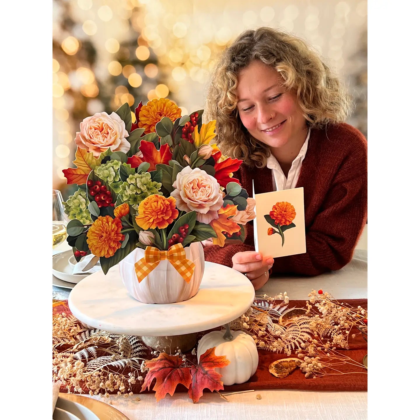 A smiling woman with curly hair sits at a table holding a small card with an orange flower illustration, while a large autumn floral arrangement in a white pumpkin-shaped vase sits on a white pedestal. The setting includes fall decorations, leaves, pumpkins, and warm blurred lights in the background.