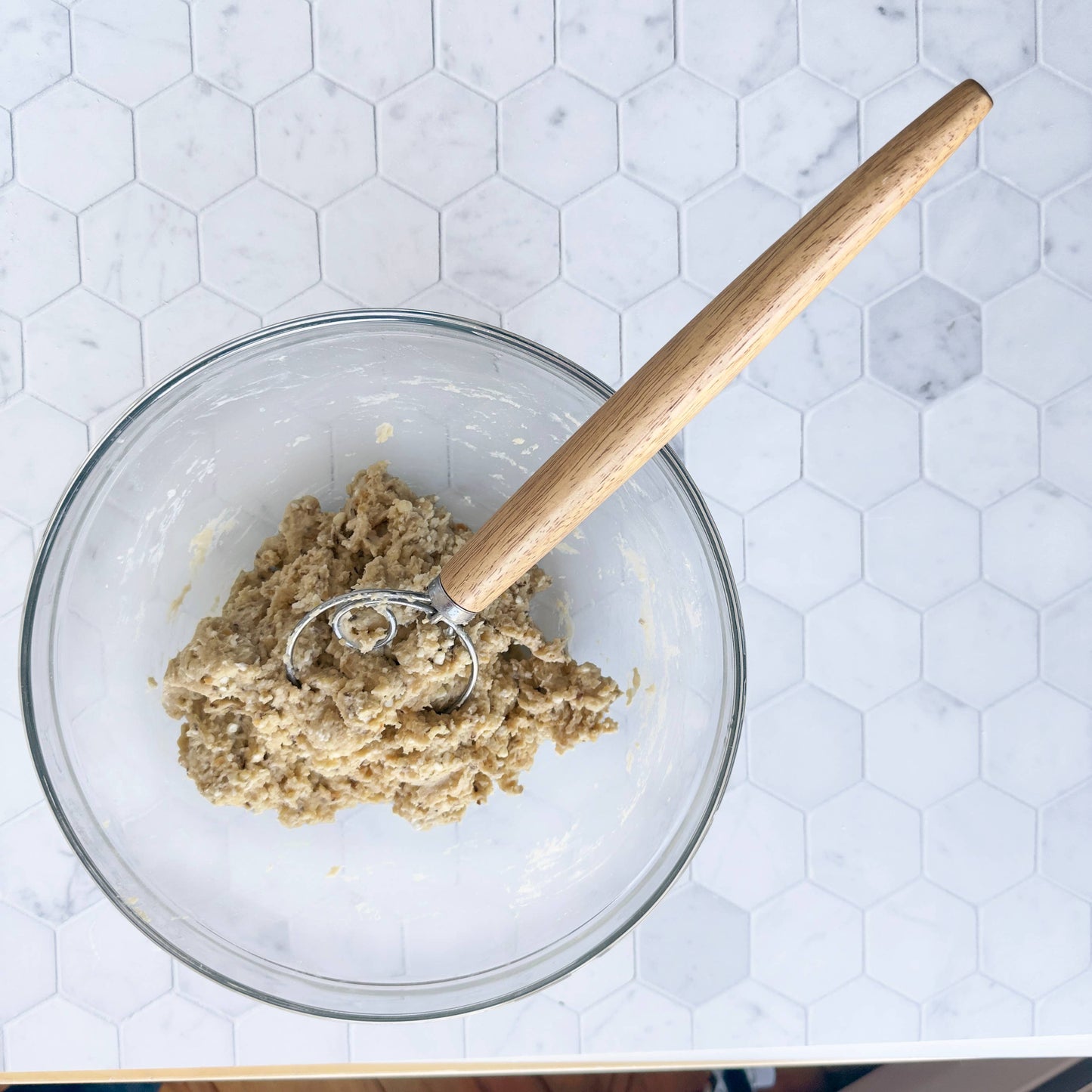 Glass bowl with cookie dough and wooden spoon on a white hexagonal tile background