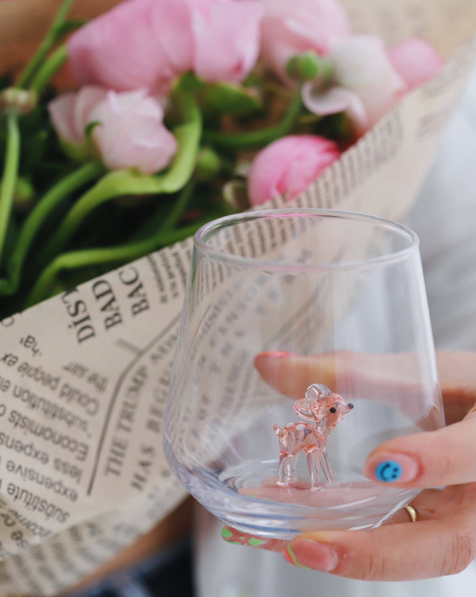 Clear glass with a small deer figurine held by a hand, with pink flowers and a newspaper in the background.