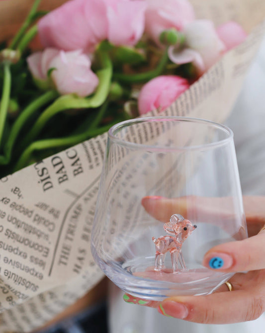 Clear glass with a small deer figurine held by a hand, with pink flowers and a newspaper in the background.