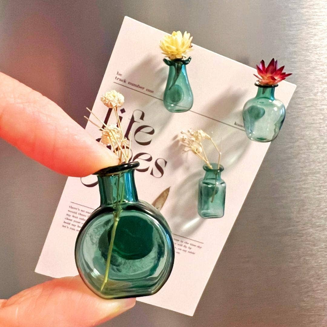 A close-up of a hand holding a small round teal glass vase with a dried flower, with three matching small glass vases attached to a white card against a metallic gray background.
