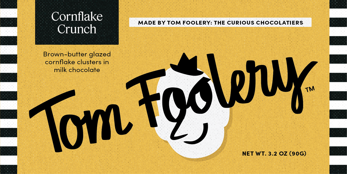 Front packaging of Tom Foolery Cornflake Crunch chocolate clusters, featuring a mustard-yellow background with black and white striped border elements, a black Cornflake Crunch label, and the Tom Foolery logo in large black script.