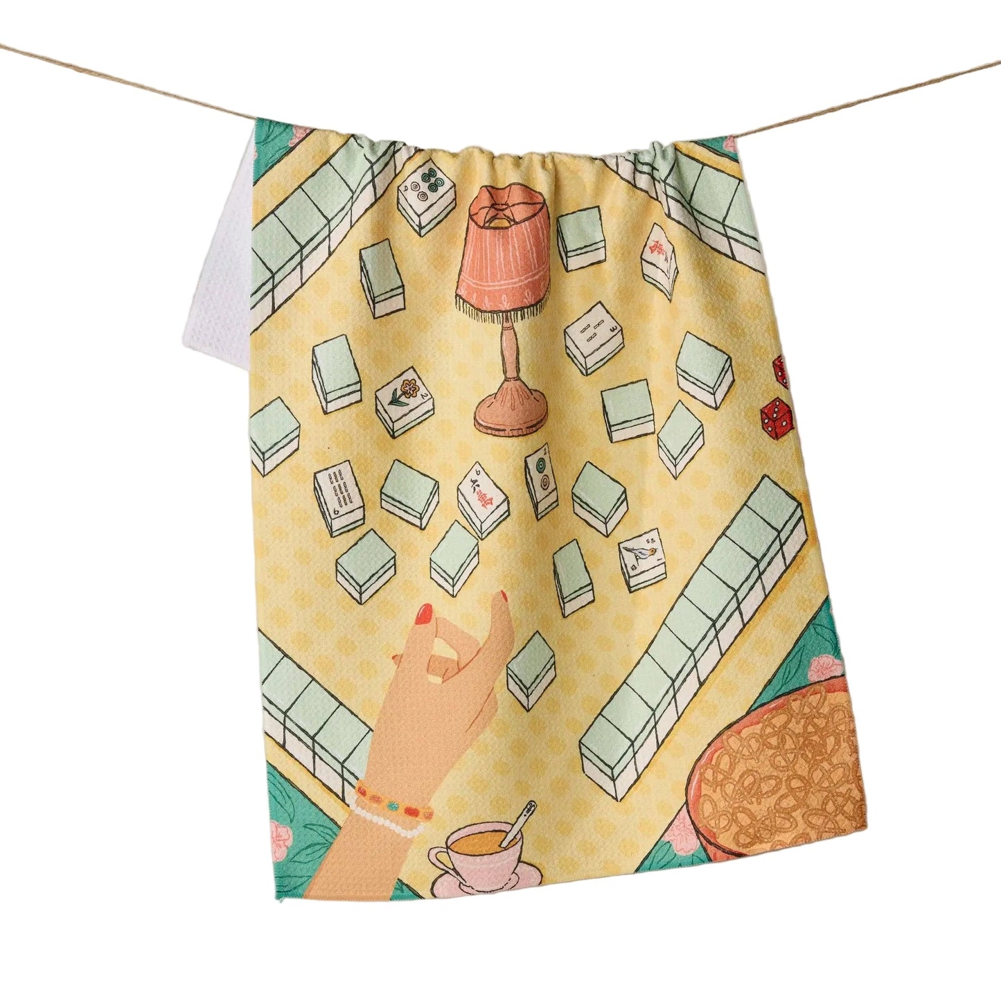 A fabric tea towel or hanging cloth printed with a stylized mahjong game scene, suspended by a string against a plain beige wall. The design shows white and green tiles, a pink lamp, a hand, and small mahjong pieces on a yellow tabletop below.