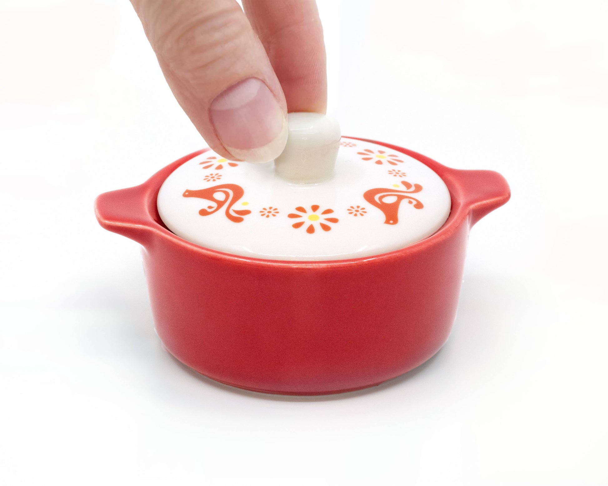 Red ceramic dish with a white lid featuring floral designs, being held by a hand.