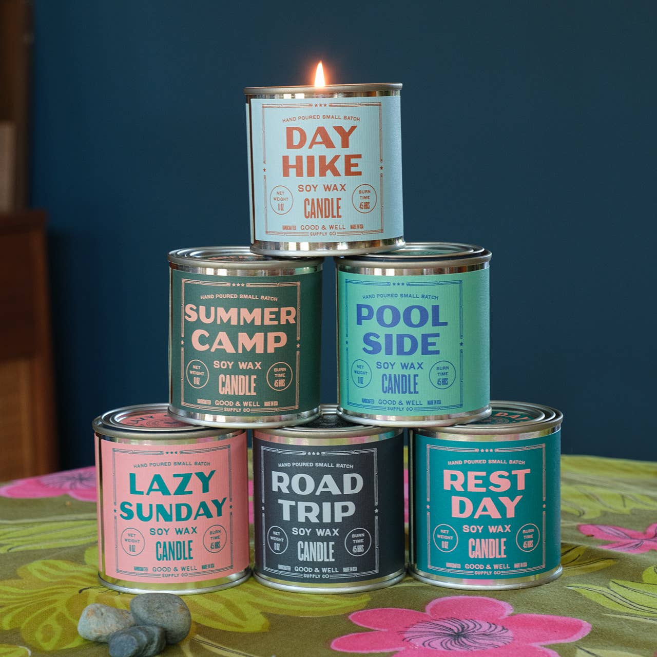 Stack of candles in metal cans with colorful labels on a floral tablecloth.