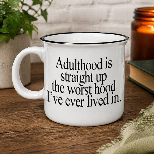 White mug with black text on a wooden surface with a plant and book in the background