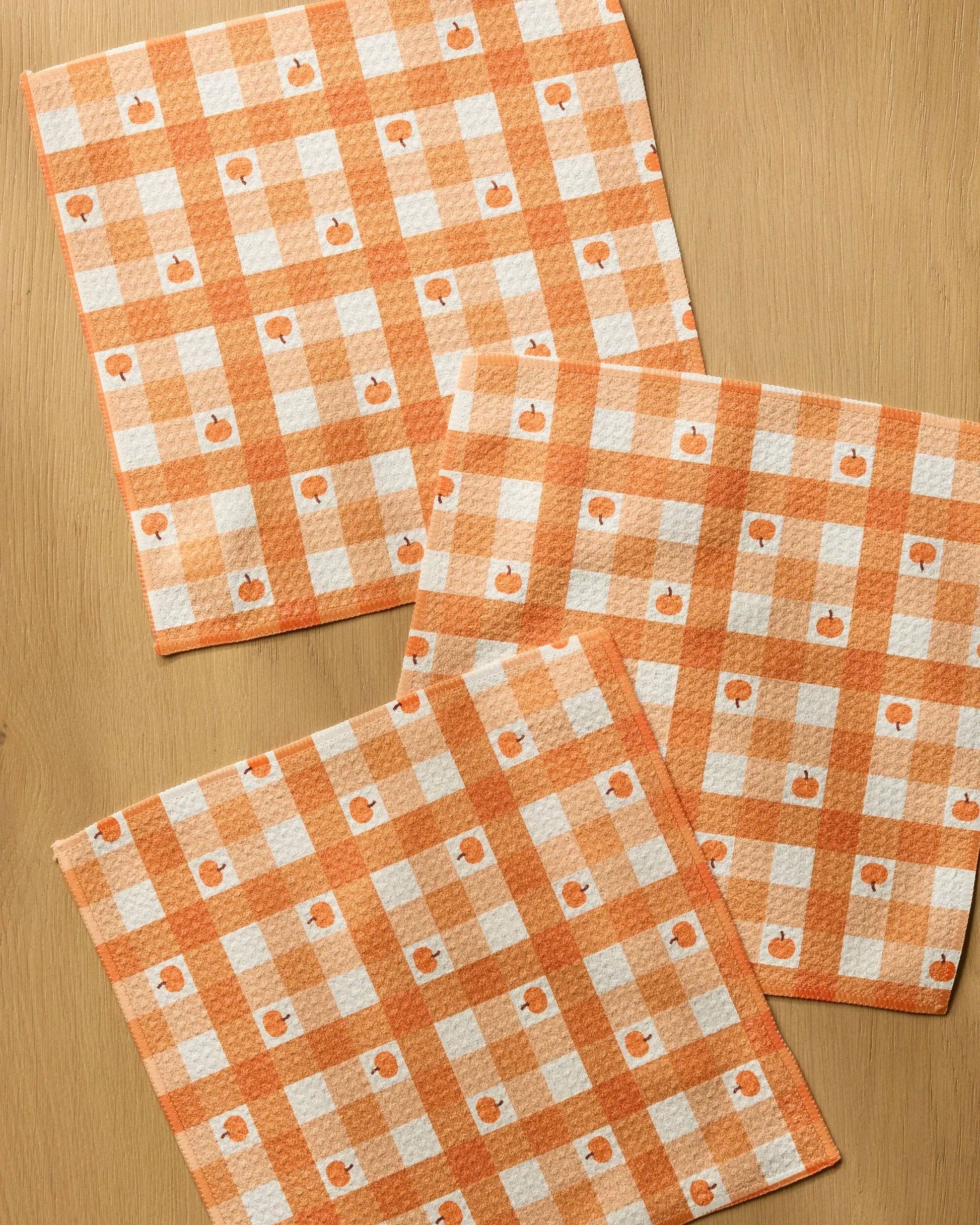 Three square kitchen towels with an orange and white gingham pattern and small pumpkin motifs, arranged on a light wood surface.