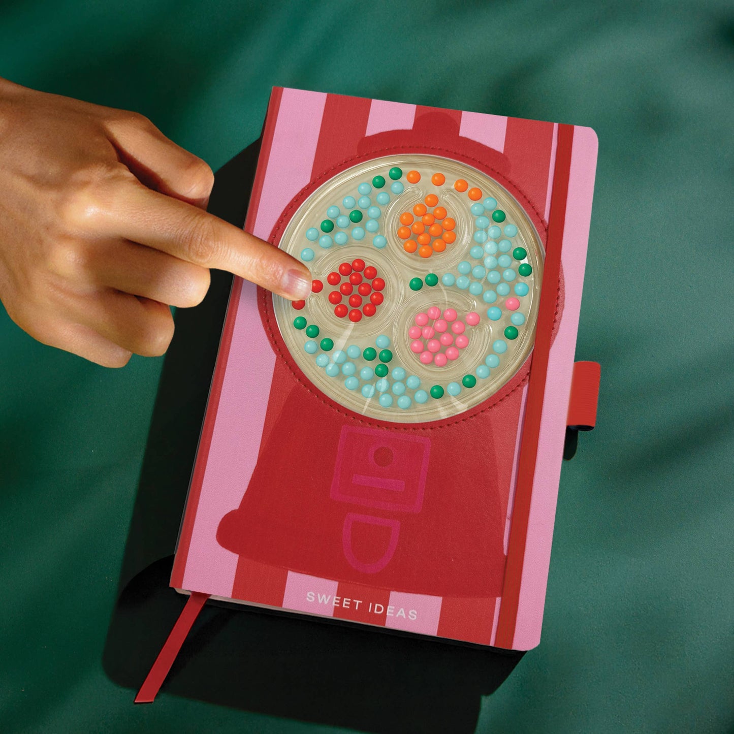 Hand poking a red and pink striped notebook with a gumball machine fidget design on a green background