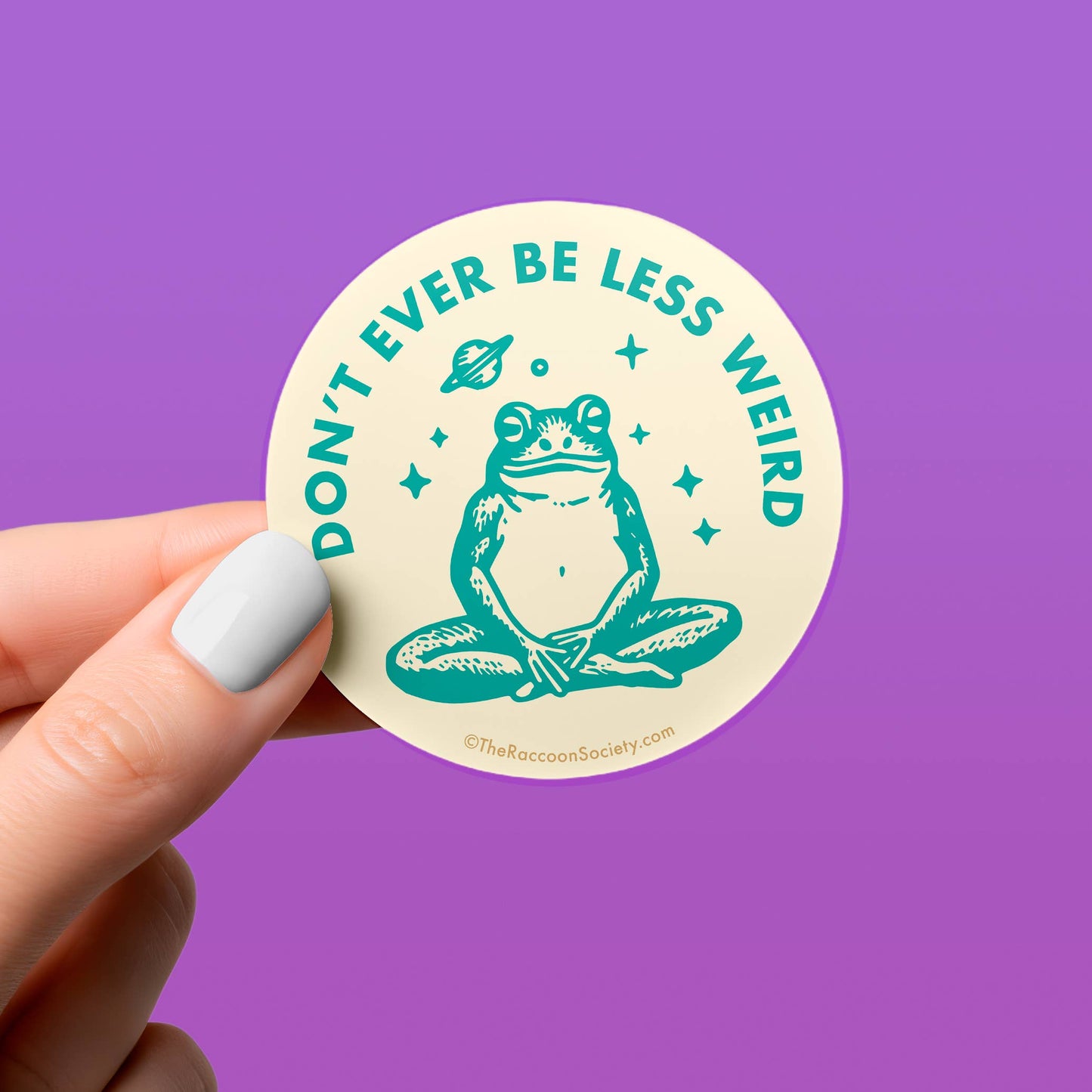 Round sticker with a frog and text 'Don't ever be less weird' held by a hand against a purple background